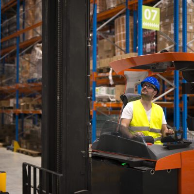 Forklift driver relocating and lifting goods in large warehouse center.